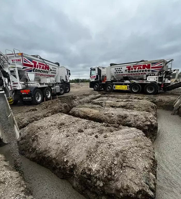 Two Titan Concrete trucks delivering ready mix concrete to a construction site. The trucks pour concrete from the volumetric mixer into a hole in the ground.