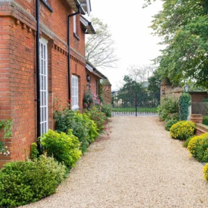 A light-coloured gravel driveway running alongside a red brick house, bordered by vibrant yellow flowers and green shrubs.
