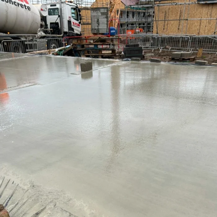 Freshly poured, wet concrete slab at a construction site with a concrete mixer truck and scaffolding in the background.