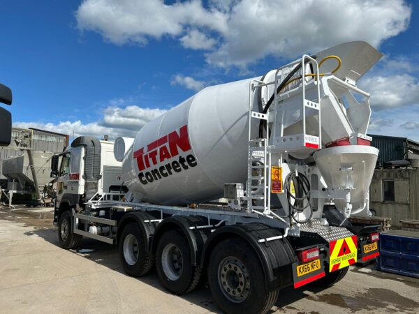 Rear-side view of a Titan Concrete cement mixer truck parked in a yard, showing the rotating drum, ladder access, and discharge chute under a bright blue sky.