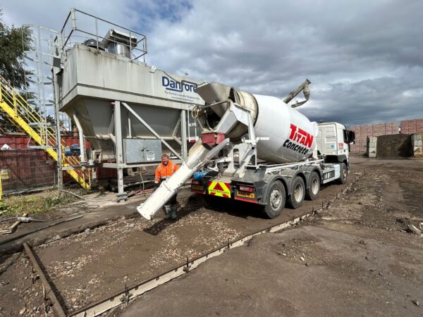 Titan Concrete mixer truck reversing toward a batching plant while a worker in high-visibility clothing stands nearby, preparing to pour concrete into a framed foundation area.